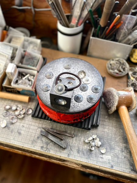 アトリエの台の上に制作途中のコンチョが並び、手仕事の空気感や数の多さが伝わる作業風景を切り取った一枚です。