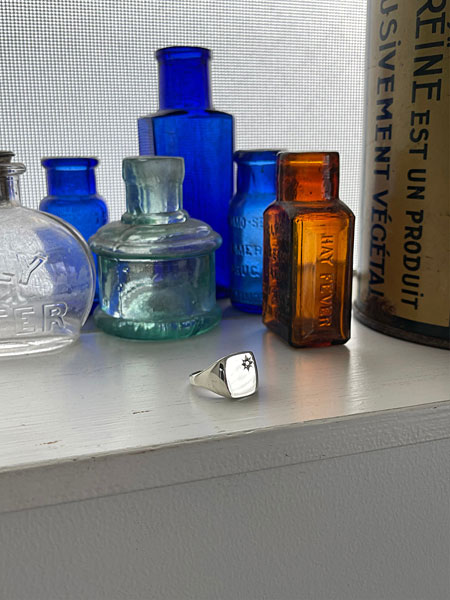 窓際でガラス瓶と一緒に並べたシグネットリングが、自然光を受けて柔らかく輝くディスプレイ画像。