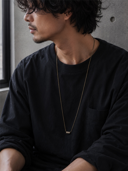 窓辺に佇むモノトーンコーデの男性の胸元に、ロングネックレスがゆったりとしたVラインを描く着用画像。