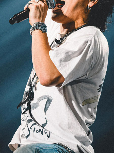 ライブで歌うミュージシャンの優里さんが、ブラックのレザーフェザーネックレスを身に着けている画像。